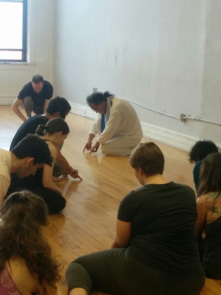 Tadashi Endo in a kneeling position, demonstrating an exercise using his fingers on the floor to make special messages. Students around him mimic his behavior. The floor is a blonde wood with light gray walls and lots of natural light. This was Outerspace in Wicker Park. 