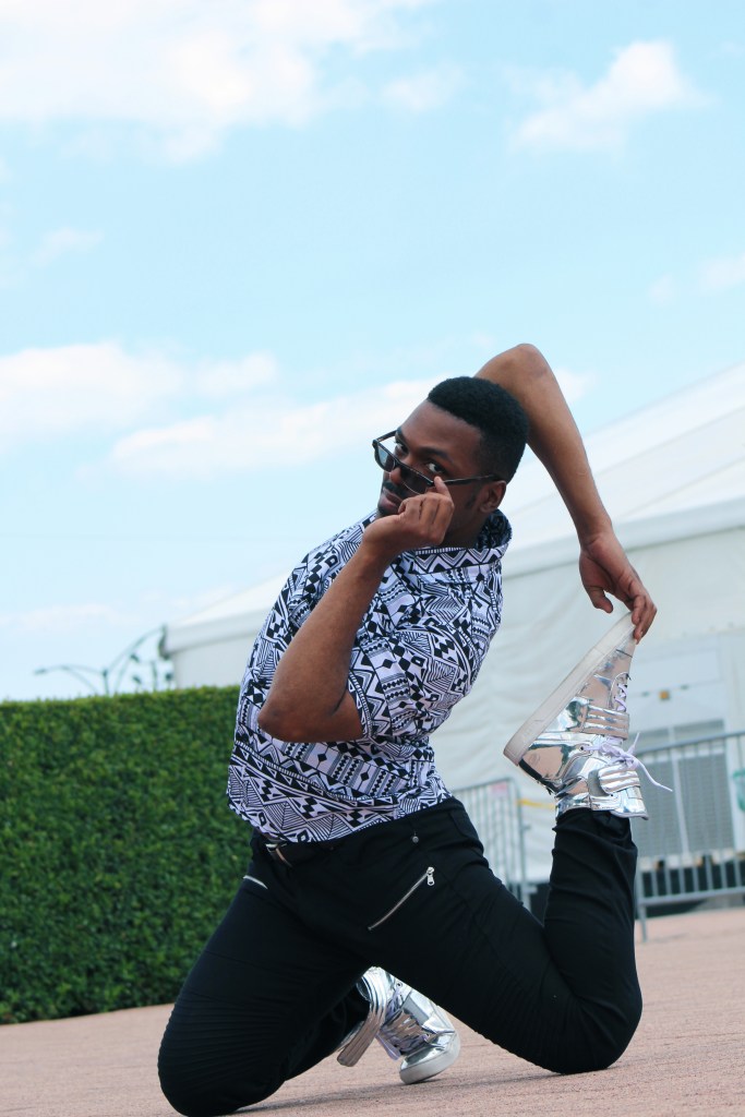 Artist Fabulous Freddie in a striking pose, kneeling, with one hand holding the opposite foot forming a loop behind their body while lowering his glasses to give us a look.