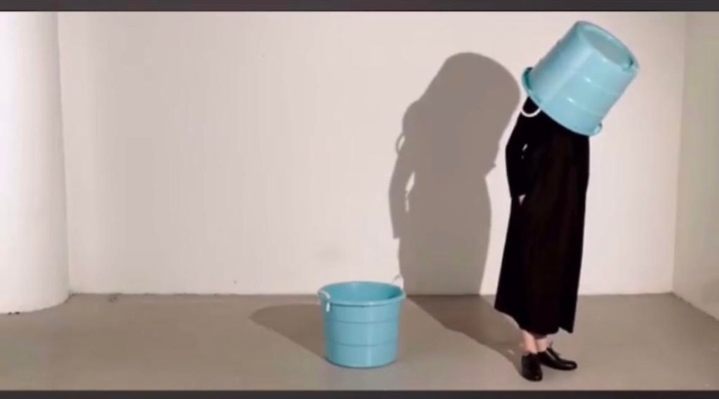 The artist Jinlu stands near the right side of the frame with a white background, black dress and shoes. She has her hand in the pocket and a giant pail upside down over her head. Another one, empty, stands beside her. Her shadow looms behind her on the wall. 