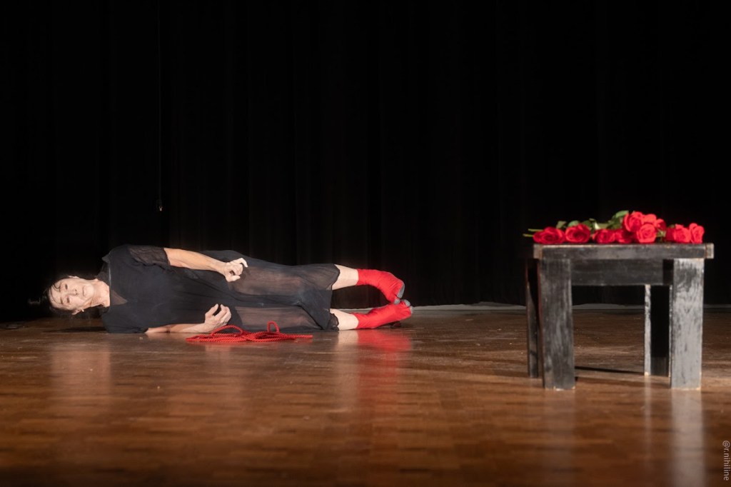 Joan Laage on the floor with arms and legs shooting out parallel in the same direction, her feet are toward a black square table with red roses on the top. The wood floor and dark black background are the only other visible cues.