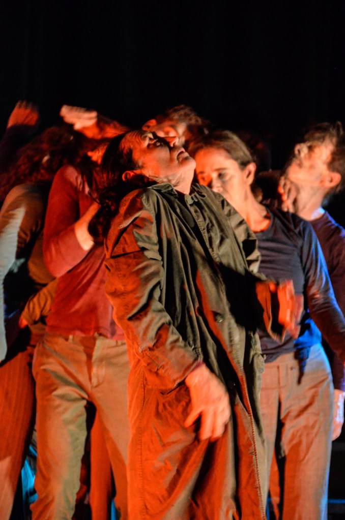 A portrait shot of group of dancers in a cluster, red lighting, Joan Laage is in the center with her face turned up in a mournful way, her hands are expressive and moving, and the faces are illuminated in these upturned, closed eyes, sensing feeling.