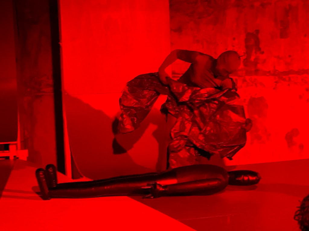 A very red photo. Sara Zalek is in a leans over an inflatable mannequin on the floor. They have a full bodysuit on so their face cannot be seen. They strike a dramatic pose holding a material which hovers over the mannequin.