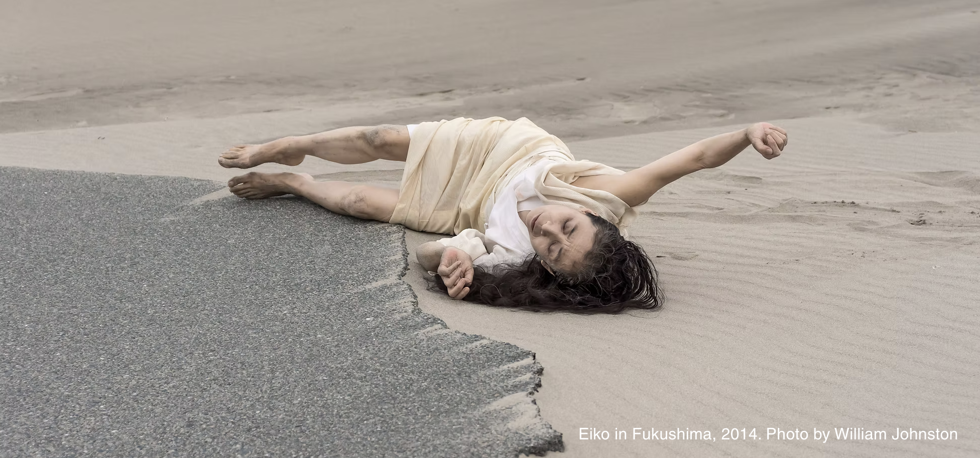 Eiko Otake in Fukushima, Japan. Photo by William Johnston. Artist Eiko pays on the edge of the water on the sand in a white dress, her face and arms to the sky.